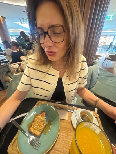 A close-up, selfie-style image captures a woman seated at an indoor café or restaurant in Senhora da Hora, Portugal. The image is rotated 90 degrees clockwise, showing the woman's face and upper body in the foreground. She wears brown-framed glasses and has light brown hair. Her attire includes a cream-colored cardigan with vertical black stripes, worn over a black top. A thin necklace is visible, and several bracelets adorn her right wrist.
On the wooden table in front of her are several items: a slice of cake, possibly a pound cake or similar, drizzled with caramel or a sweet sauce, presented on a teal plate with a fork. Next to it, there's a white cup containing dark coffee (likely an espresso) on a white saucer. A large glass jug filled with a bright yellow beverage, consistent with orange juice, is also present.
The background reveals a modern and comfortable interior. There's a soft green upholstered bench or couch, and walls feature horizontal wooden paneling. Other patrons can be seen seated in the mid-ground, and plants contribute to the decor. Bright indoor lighting illuminates the scene. Faint, rotated text that appears to be "ABBA" or "ARBA" is visible on a pillar in the background, along with a green exit sign. The overall scene suggests a relaxed daytime meal or snack.