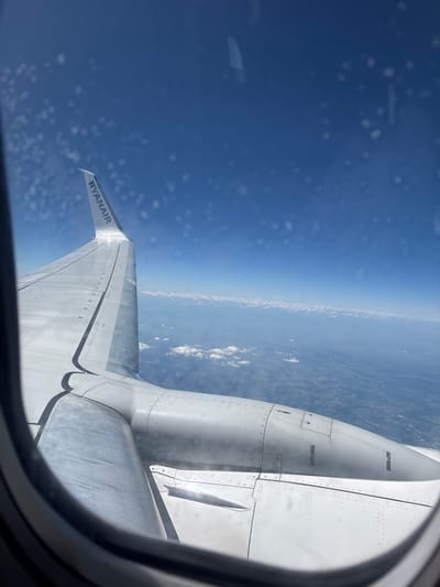 The image is an aerial view from an airplane window, looking out at the wing and the landscape below. The scene is set during the day, with bright sunlight illuminating the airplane's wing, which is the primary object in the foreground. The wing is metallic and appears to be made of segmented panels, with visible rivets. The "RYANAIR" logo is clearly visible in dark text on the edge of the wing.

In the mid-ground, the curvature of the Earth is visible, with a clear distinction between the deep blue of the sky and the hazy horizon where the atmosphere thins. The land below is a mosaic of green and brown, suggesting a rural or semi-urban area with some patches of white clouds scattered across the landscape. The overall impression is one of travel and distance.

The image is taken from inside an airplane, evidenced by the circular window frame visible in the lower right portion of the image. The window itself has some water droplets or condensation on it, creating a slightly blurred effect and some white specks in the sky. The lighting suggests it is daytime, possibly late morning or early afternoon, given the angle of the sun. There are no people visible in the image. The overall mood is one of peaceful observation of the world from above.

The provided location context "Majdalena, Czechia" suggests the flight path is over this region.
