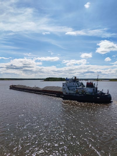 A tugboat and barge traverse a body of water on a sunny day with a blue sky punctuated by cumulus clouds. The barge appears to be laden with a dark, granular material. In the distance, a shoreline with trees is visible. The water reflects sunlight, creating a shimmering effect. The city of Novodevich'e, Russia is not readily identifiable from this image.