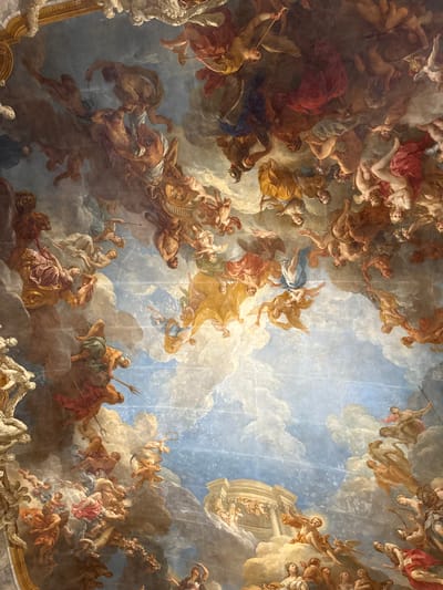 This image depicts a highly detailed Baroque-style ceiling fresco, identified as "The Apotheosis of Hercules" by François Lemoyne, located in the Hercules Salon of the Château de Versailles, Le Chesnay-Rocquencourt, France.

The painting presents a dense, multi-figure mythological scene spanning a vast celestial realm composed of a blue sky and voluminous white and grey clouds. Numerous human and anthropomorphic figures, male and female, are depicted in various states of motion and repose, occupying the entire painted surface. Most figures are nude or draped in flowing classical garments of red, gold, blue, white, and earthy tones.

Key elements observed:
*   **Figures:** Over a hundred individuals are present, including powerful male figures (one wielding a trident, consistent with Neptune, in the lower-left), female deities, winged putti, and other mythological beings. A central, muscular male figure, often identified as Hercules, is prominent, appearing to ascend or be lifted amidst the clouds. Many figures bear symbolic attributes such as trumpets, scepters, or bows.
*   **Actions and Interactions:** Figures are shown flying, floating, rising, or resting on clouds. Interactions vary from dynamic struggles and ascensions to celebratory gestures and contemplative gazes. Several figures appear to be converging towards a bright area or a specific architectural element.
*   **Objects/Structures:** In the lower-right quadrant, a classical temple-like structure with columns is depicted on clouds, bathed in light, possibly representing Olympus or a heavenly destination. An ornate gold architectural trim is partially visible along the top edge of the frame, indicating the boundary of the fresco.
*   **Environment:** The background consists of an illusionistic sky, ranging from deep blue to lighter hues, filled with dramatically lit and shadowed clouds that create an impression of immense depth and a limitless heavenly expanse.