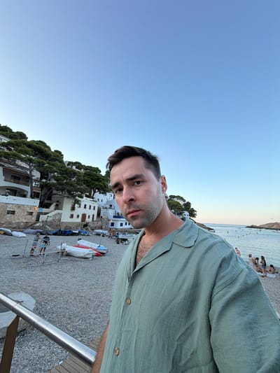 The image captures a selfie of a man on a pebble beach in the town of Begur, Spain. The man, wearing a light green button-down shirt, is positioned in the foreground, with his body taking up a significant portion of the right side of the frame. He has short, dark hair and is looking directly at the camera with a neutral expression.
Behind him, the beach is lined with several small boats, some covered with tarps. Further in the background, white buildings with red-tiled roofs, characteristic of Mediterranean architecture, are nestled on a hillside covered with green trees. The sea is visible on the right side of the image, with calm water and a few buoys in the distance. Several people are scattered on the beach and in the water, engaged in various activities like sunbathing and swimming. The sky is clear and blue, suggesting it is daytime, likely late afternoon or early evening due to the soft lighting. The overall scene conveys a relaxed, coastal atmosphere.