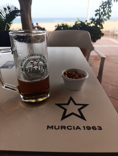 The image shows a close-up of a table at an outdoor cafe with a beach and ocean in the background. On the table is a half-full beer mug with the logo "Estrella de Levante" and a small bowl of nuts. In the background, there is a sandy beach, the ocean, and a few people walking. The table itself has a black star logo and the text "MURCIA 1963". The location is likely a cafe or restaurant along the coast of Águilas, Spain, given the context of the beer brand and the beach setting. The lighting suggests it is daytime, possibly overcast.