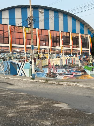 The scene depicts a vibrant streetscape in General Villamil Playas, Ecuador, centered on a two-story building with a distinctive architectural style. The upper portion of the building features an arched roof with alternating light blue and white vertical stripes. Below the roof, the facade is composed of multiple large, rectangular windows with a reddish-orange tint, reflecting either the sky or internal lighting. Below these windows, a white metal railing system indicates an elevated walkway or balcony. The lower level of the building or a retaining wall extending from it is adorned with a colorful, detailed mural. The left section of the mural displays abstract geometric patterns in shades of blue, white, and black. The right section of the mural depicts a detailed maritime scene with several sailboats with reddish sails and motorboats on blue water, set against a background of a city skyline with multiple high-rise buildings. The words "PLAYAS DE VILLAMIL" are visible on the mural, and "A ELVIS PLAVAS" is written on one of the motorboats. One individual, dressed in dark clothing, is partially visible sitting or leaning on the white railing system above the mural. In the foreground, a heavily loaded utility pole with numerous black electrical wires and a street lamp stands centrally. The pole is painted in light blue and white stripes at its base. The ground consists of a paved road in the immediate foreground, with a dirt and gravel shoulder. A low concrete curb separates the road from the mural structure. The sky above is clear and bright.