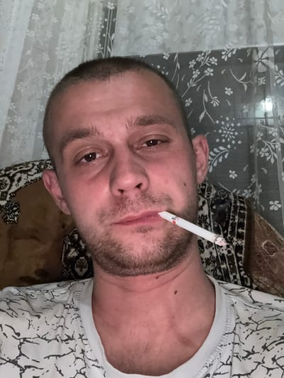 A man is holding a lit cigarette in his mouth while looking directly at the camera. He has short, dark hair and stubble on his face. He is wearing a white t-shirt with a black abstract pattern. The background consists of a curtain with a floral pattern. The setting appears to be indoors, likely a residential room. The image is a close-up, focusing on the man's face and upper body. There is no indication of the specific location within Астроміцкі сельскі Савет, Belarus.