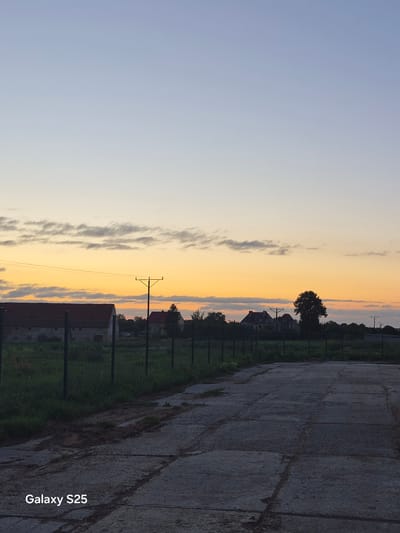 The image depicts a rural landscape at dusk. The sky is a gradient of pale blue and soft orange, with wispy clouds scattered across the horizon. In the middle ground, there are several buildings with red roofs, possibly farm structures or houses, partially obscured by trees and a fence. A power line stretches across the frame, supported by a utility pole. The foreground features a rough, concrete surface, perhaps a road or a paved area, with patches of grass and dirt. The overall impression is one of a quiet, rural setting as day transitions into night. The location is identified as Pątnów, Poland, indicated by the text "Galaxy S25" at the bottom left corner of the image. There are no people or animals visible in the scene.