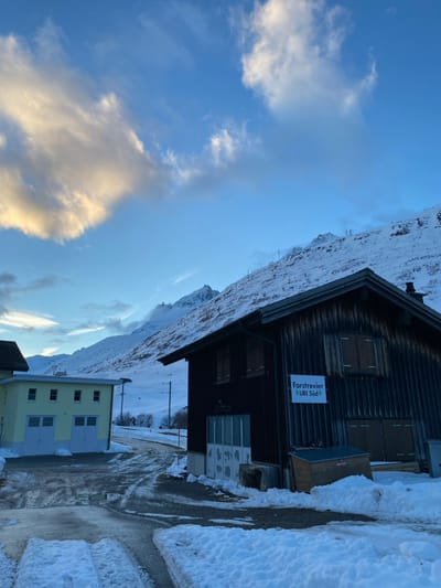 The scene depicts a snow-covered mountainous landscape in Hospental, Switzerland, under a clear blue sky with scattered white and light orange-hued clouds. In the foreground-right, a two-story dark brown wooden building with a gabled roof is present. A white sign affixed to its side reads "Forstrevier URI Süd" (Forest District URI South). Multiple windows on this building are covered by closed wooden shutters. A dark, possibly wooden, storage unit or box is positioned at ground level next to the building, partially obscured by snow. To the left, a light yellow two-story building with multiple rectangular windows and two large, lighter-colored garage doors is visible. The ground between and in front of the buildings is covered in snow, with a partially cleared or melted asphalt/gravel road showing tire tracks. The background is dominated by a large, snow-covered mountain range, with utility poles or similar structures faintly visible on its lower slopes. No individuals, actions, or direct interactions are depicted.