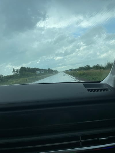 The image, taken from inside a vehicle, shows a wet road stretching into the distance under a cloudy sky. Raindrops are visible on the windshield, distorting the view of the landscape which consists of trees and fields. The dashboard of the car is partially visible in the foreground, with an air vent and the "AIRBAG" warning light. A white sign is visible on the left side of the road. Based on the provided metadata, the location is Жаңаауыл, Kazakhstan.