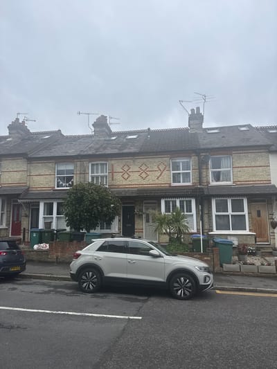 The image shows a row of terraced houses in Watford, United Kingdom. The houses are made of yellow brick with red brick detailing. They have pitched roofs with dormer windows and chimneys. The houses are numbered 16, 18, and 20. House number 18 has the year "1889" displayed in brickwork above the first-floor windows.
In front of the houses, a grey car is parked on the street. Several green and blue wheelie bins are lined up along the pavement. A large green tree is planted in front of house number 18. The sky is overcast and grey.