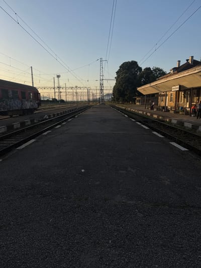 The image depicts a train station platform in Kaspichan, Bulgaria, under a clear, pale sky, suggesting either sunrise or sunset. The perspective is from the edge of a dark asphalt platform, looking down a set of train tracks that recede into the distance.

On the left, a weathered red train car, covered in graffiti, is parked on a parallel track. Overhead, a complex network of electrical wires and supporting pylons crisscrosses the sky, typical of a train station.

To the right, a low-slung building, the station house, features a covered waiting area with benches and support columns. Above the entrance, signage is visible, appearing to read "КАСПИЧАН" (Kaspichan) in Cyrillic script. A few individuals are present near the building, some appearing to be waiting or standing. One person is seated on a bench under the awning, while two others stand near the entrance.

The platform itself is sparsely populated, with only a few scattered small objects visible on the asphalt. The environment appears to be a functional, albeit somewhat dated, railway facility. The overall impression is one of quiet activity at a regional train station during a period of transitional light.