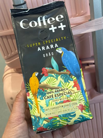 A human left hand holds a "Coffee ++ Super Specialty - Arara" coffee bag, angled slightly upwards and to the right. The upper portion of the bag is matte black with white "Coffee ++" branding and yellow "SUPER SPECIALTY - ARARA GRÃO" text. A circular degassing valve is centrally located below the brand name. The lower half of the bag features a vibrant tropical illustration depicting a yellow macaw and a blue macaw perched on a brown branch, surrounded by green leaves and red hibiscus-like flowers on a pale yellow background. Product specifications include "100% ARÁBICA", "CAFÉ ESPECIAL", "TORRA MÉDIA" (Medium Roast), "CAFÉ TORRADO EM GRÃO" (Roasted Coffee in Bean), "250g", "WHOLE BEAN", and "NET WT 8.8OZ". In the background, out of focus, are elements of a light pink ribbed hard-shell suitcase, including a pink fabric zipper and a gray metallic hexagonal luggage handle. The environment suggests an indoor travel setting. The image itself does not contain verifiable information to pinpoint the location specifically within the city of Guarulhos, Brazil.