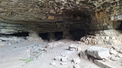 This is a photograph of the entrance to a cave in Milanovo, Bulgaria. The cave is wide and shallow, with a rocky, uneven floor littered with fallen rocks and some patches of green vegetation. The ceiling of the cave is composed of layered rock formations, with some areas showing signs of erosion and mineral deposits. The walls of the cave are also made of layered rock, with some sections appearing rough and others smoother. There are several openings or smaller caves visible within the larger cave entrance. Graffiti is visible on some of the rock surfaces, particularly on the right side of the image. The overall impression is of a natural, rugged environment.
