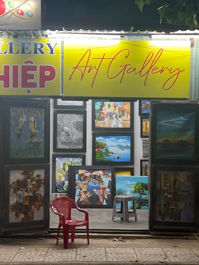 The media file displays an art gallery entrance at what appears to be daytime, with a bright yellow sign that reads "ART GALLERY" in red cursive script. Below this, another sign in blue and red lettering says "GALLERY HIỆP". The gallery is filled with numerous framed paintings, displayed both inside and outside the entrance. The paintings vary in style and subject matter, including landscapes, abstract art, and depictions of village scenes. Some are large and prominent, while others are smaller and arranged in clusters.

A bright red plastic chair sits outside the gallery, suggesting a waiting area or an informal display space. The ground beside the gallery is paved with decorative tiles, common in many Southeast Asian countries, and there is a hint of greenery from some plants above the entrance. The overall impression is of a bustling, visually rich environment, typical of a tourist-frequented area like Hoi An, Vietnam, where art and crafts are a significant part of the local economy. The lighting suggests it's daytime, and the weather appears to be clear. There are no people actively engaged in activities within the visible frame, but the display of art implies it is open for business.
