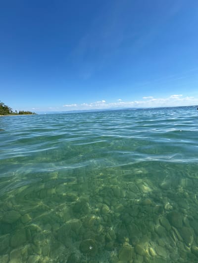 The image depicts a serene, sun-drenched scene of clear, shallow water with a rocky bottom, likely a lake or a calm sea. The water is a vibrant green, revealing smooth, rounded pebbles beneath its surface, which are illuminated by the sunlight filtering through the rippling waves. The waves themselves create a shimmering, distorted effect on the underwater landscape.
The water extends to the horizon, where it meets a bright blue sky dotted with scattered, wispy white clouds. The transition from water to sky is sharp, with a thin strip of green vegetation visible along the distant shoreline. This vegetation appears to be trees or bushes, suggesting a natural, perhaps undeveloped, coastal area. The overall impression is one of a peaceful, idyllic day.
The time of day is clearly daytime, given the bright sunlight and clear blue sky. The weather appears to be fair and warm. The location context provided places this scene in Hagnau am Bodensee, Germany, which is a town on the shores of Lake Constance. This aligns with the visual cues of clear, shallow water and a lakeside environment. There is no visible text in the image. The scene evokes feelings of tranquility and natural beauty.