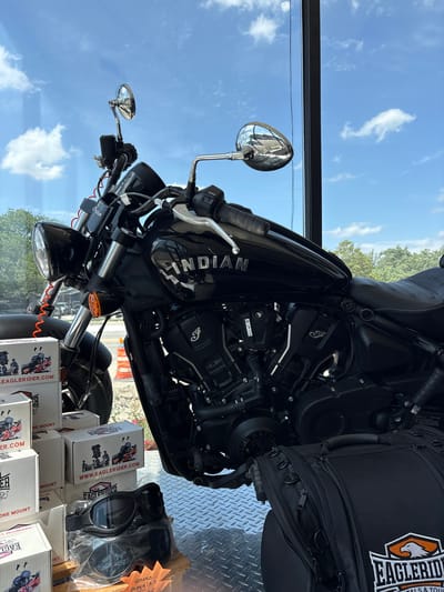 A black Indian motorcycle is displayed in a showroom, likely at Eagle Rider, a motorcycle rental and tour company, given the branding on the boxes and bags. The motorcycle is parked indoors, with a large window in the background revealing a bright blue sky with scattered white clouds, and trees and buildings visible outside. Several cardboard boxes, some with branding and images of motorcycles, are stacked to the left of the motorcycle. A pair of black goggles sits on top of one of the boxes. A black motorcycle bag with the Eagle Rider logo is placed on the floor to the right of the motorcycle. The scene appears to be a sunny day, and the motorcycle is clean and in good condition, suggesting it is for sale or rent. The overall impression is of a well-lit and organized display aimed at motorcycle enthusiasts.