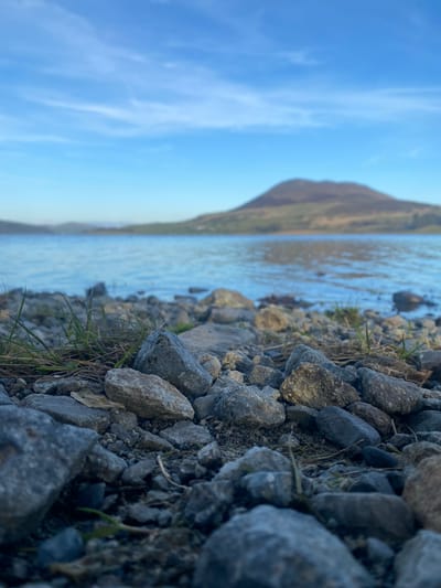 The image captures a rocky shoreline with a body of water extending towards a blurred mountainous landscape under a clear blue sky. The foreground is dominated by a variety of grey rocks, interspersed with patches of green grass. A mountain rises in the background, its peak obscured by the angle and distance. This scene depicts a portion of Bala Lake (Llyn Tegid) in Bala, North Wales, United Kingdom. The camera is positioned low to the ground, emphasizing the texture of the rocks and the proximity of the water. There is no human presence visible in the scene.