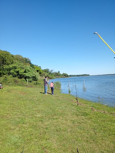 The image depicts a scene at the edge of a body of water, likely a river, in Corrientes, Argentina. Two people stand on a grassy bank near the water's edge. One person is adjusting fishing equipment, while the other is looking at the water. Several fishing rods are standing near the shoreline. The background features trees and a clear blue sky.