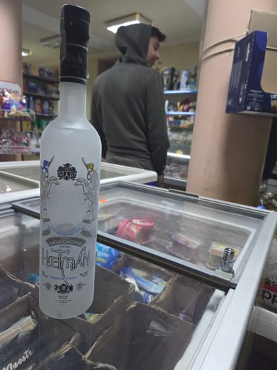 The image shows a bottle of Hetman premium vodka lying horizontally on a glass surface. The bottle is white with black and silver labeling featuring Ukrainian text. The text includes the words "Hetman", "Premium Vodka", "Sagaidachny", and "Made in Ukraine." In the background, a person wearing a grey hooded sweatshirt is visible near a refrigerated display case stocked with various items. Shelves with merchandise are visible in the blurred background further into the store. A Lavazza box is visible on the upper left. The location in the city of Pripek, Bulgaria is inferred from other sources and cannot be directly determined from the image itself.
