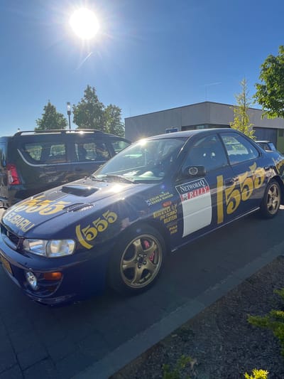 A blue Subaru Impreza rally car replica is prominently featured, parked in what appears to be a paved lot in Reckange-sur-Mess, Luxembourg, on a bright, sunny day. The car is adorned with yellow "555" decals on its side and front, along with various sponsor and rally-themed stickers. Visible text includes "NETWORKO RALLY," "X COLIN MCRAE," "X DEREK RINGER," "PIRELLI," "IHI TURBO," "BILSTEIN," "KENWOOD," "PIAA," and "NGK." Part of the "SUBARU" branding is also visible. The car has gold-colored wheels with red brake calipers.
Behind the Subaru, a darker SUV or van is parked, with "LOGAN" visible on its rear. The setting includes buildings and green trees in the background. The sky is clear blue, with the bright sun creating a strong lens flare and reflections on the vehicles. A distinct white contrail from an aircraft is visible high in the sky. No specific activity or event is taking place beyond the cars being parked.