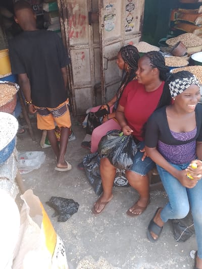This image captures a vibrant market scene in Mgbuoshimini, Nigeria, under bright daylight conditions. The setting appears to be an outdoor market stall or the entrance of a shop, characterized by a concrete ground and a metal shuttered door in the background.

The subjects include a young man and three women. The young man, with short dark hair, is seen from behind, wearing a black t-shirt and orange and brown patterned shorts, with light-colored flip-flops. He stands near a large metal basin filled with light-colored grains, possibly groundnuts or peanuts. To his right, three women are seated. The woman closest to the camera has braided hair and wears a distinctive black and white patterned headband, a black top over a purple and white striped tank top, and blue jeans. She is smiling broadly, holding a yellow object (possibly a drink bottle). Next to her, a woman with braided hair wears a red t-shirt and dark denim shorts, holding a black plastic bag on her lap. Another woman, also with braided hair, is partially visible behind them.

The scene is bustling with market elements. Large basins filled with what appear to be grains are visible on both sides of the image. Numerous sacks and bags, some white and some brown, are stacked in the background, suggesting various goods for sale. A black plastic bag rests on the ground near the seated women, and another large white bag with "AR" and an emblem is visible on the floor. The metal shuttered door in the background features red graffiti-like markings and several posters or stickers, though most text is illegible, except for partially visible words like "WINNER" and a date like "2017". The overall activity suggests people are either shopping, selling, or resting at a market stall. The bright light and the smiling woman in the foreground indicate a pleasant, probably warm, day.