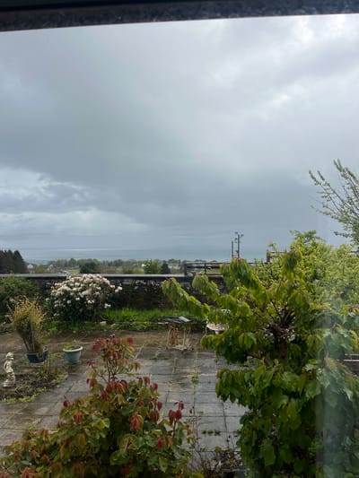 The image depicts an exterior scene, likely a view from inside a building in Golspie, United Kingdom. Overcast skies dominate the upper portion of the image. A body of water, possibly the sea, is visible in the middle distance with a thin strip of land. Trees and shrubs are present in the midground, and a garden or patio area is in the foreground, including flowers, paving stones, and potted plants. The presence of power lines suggests a residential area.