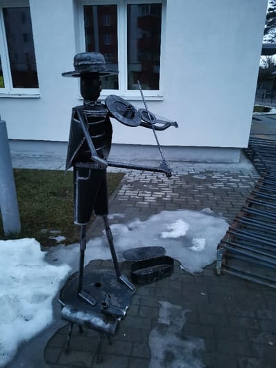 An outdoor scene in Masty, Belarus, features a dark grey metal sculpture depicting a figure playing a violin. The sculpture is constructed from welded metal components, including a wide-brimmed hat, an elongated head, a body resembling a jacket, thin legs, and outstretched arms holding a violin and bow. It stands on a circular metal base with three short legs. To the right of the sculpture's base, an open, dark rectangular metal container rests on the pavement. The ground beneath the sculpture and extending to the right is paved with grey rectangular interlocking pavers, showing patches of melting snow and ice. To the left of the sculpture, a patch of green grass with remnants of snow is visible, alongside a partial view of a vertical grey pole. In the background, a light-colored building with two large windows reflects a multi-story residential building and bare trees. On the far right, a dark, slatted metal structure is partially visible. The lighting is diffused, indicating overcast conditions or twilight.
