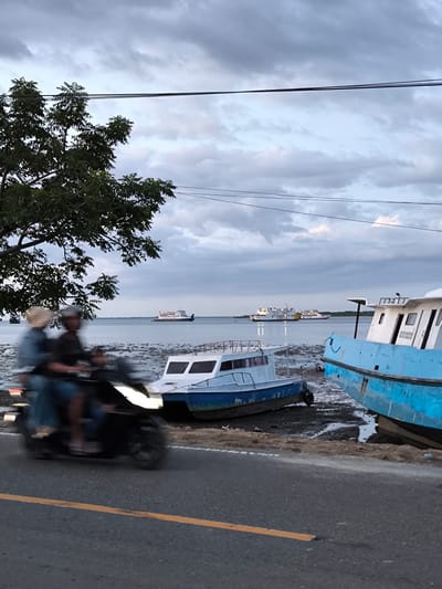 The image captures a coastal scene in Upt Tambak Sari, Indonesia, under an overcast sky. Several boats are beached on the muddy shore, with a blue and white vessel prominently featured. In the distance, two larger ferries are visible on the water. A tree stands to the left side of the frame, partially obscuring the view. A scooter with two passengers is passing by on the road in the foreground. The road is paved with a yellow line. Overhead wires are present.