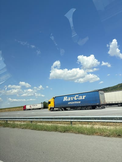 The image shows a highway scene on a clear, sunny day. The sky is a vibrant blue with scattered white clouds. In the foreground, a highway with two lanes is visible, separated by a guardrail. Beyond the highway, a field of dry grass and some vegetation lines the road.
Several trucks are on the highway. In the middle ground, a large blue semi-trailer truck with "RavCar SPEDITION" written on its side is prominently featured. To the left of the blue truck, a white semi-trailer truck with red accents and the logo "KAYA LOGISTIK" is visible. Further in the background, another white semi-trailer truck is partially visible. The trucks appear to be in motion. The landscape behind the trucks consists of rolling hills covered in vegetation.
While the prompt mentions the city of Nedyalsko, Bulgaria, there are no specific visual cues within the image that confirm this location. The scene depicts a generic highway environment that could be found in many regions. There are no people visible in the image.