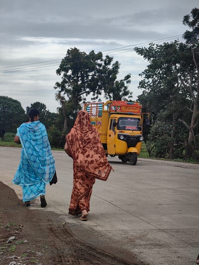 In Walsawangi, India, two women walk along a paved road next to a grassy shoulder. The woman on the left wears a blue and white patterned sari and walks on the road. The woman on the right wears a peach-colored sari with a brown floral pattern and walks on the road. Behind the women, a yellow auto-rickshaw with a covered cargo area is driving on the road. The auto-rickshaw has text on its front and sides. Trees line the right side of the road, and a cloudy sky is visible above.