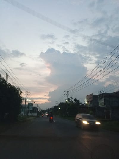 The image depicts a street scene at dusk, possibly in Sagoe, Indonesia, characterized by a twilight sky with scattered clouds. A road runs through the center of the frame, with vehicles visible on it. In the foreground, a dark-colored car with its headlights on is blurred due to motion. Further down the road, a motorcycle with its tail light illuminated is also in motion. The left side of the road features trees and utility poles with numerous overhead wires, some of which crisscross the sky. On the right side of the road, there are buildings and more utility poles. The overall lighting suggests a low-light environment, typical of sunset or twilight.