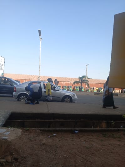 The image depicts a roadside scene in Ushapa, Nigeria, under a clear blue sky. In the midground, a silver four-door Opel Astra G sedan is parked on the edge of an asphalt road and concrete sidewalk. Its front passenger side tire is visibly deflated. One individual, dressed in a light blue top and dark long skirt, is bent over near the deflated tire, appearing to attend to it. Another individual, wearing a yellow long-sleeved top and light-colored pants, stands next to the open front passenger door, observing. To the far left, part of a dark-colored vehicle is visible. On the far right, a person in a patterned top and dark skirt walks from right to left.
In the background, a row of green and yellow motorized tricycles is parked in an open area. Behind them stands a long building with a reddish-brown tiled roof. A tall white solar-powered street light pole rises prominently in the mid-left, and a utility pole with wires is visible further right. A single palm tree is also present. A partially visible sign on the left indicates "FEDERAL STATE GOVERNMENT" and "UBU." The foreground consists of dirt ground and a concrete sidewalk with a drainage channel.