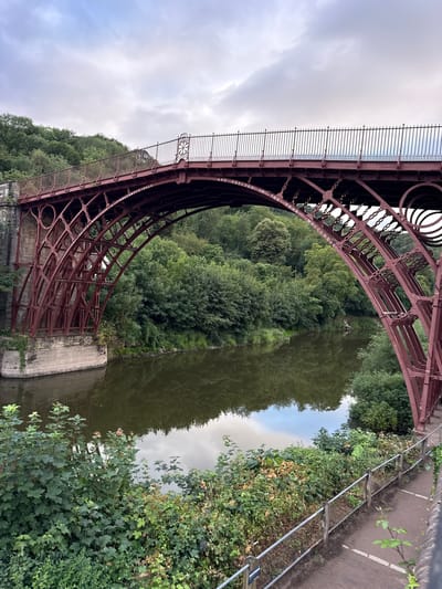 The image shows the Iron Bridge, a cast iron arch bridge that spans the River Severn in Ironbridge, Shropshire, United Kingdom. The bridge is painted a deep red and is characterized by its intricate lattice-like structure. The river below is calm and reflects the surrounding lush green trees and the overcast sky. The banks of the river are covered in dense foliage, and a paved walkway with a metal railing can be seen in the foreground. The sky is cloudy, suggesting a temperate day. No people are visible in the frame. The bridge appears to be well-maintained, with some moss and greenery growing on its stone supports. The inscription "THE BRIDGE WAS CAST AT COALBROOK-DALE AND ERECTED IN THE YEAR MDCCLXXIX" is visible on the arch of the bridge, indicating its construction date of 1779.