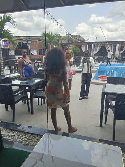 The image shows a group of people standing in a restaurant in Ujevwu, Nigeria, located at latitude: 5.472, longitude: 5.817. The restaurant has a pool in the back. The women in the image appear to be socialising while a man stands at the edge of the pool. One of the women is wearing a floral dress, while the other two are wearing casual outfits. The restaurant is decorated with furniture, tables, and a small bar, and there are plants in the background. The environment appears to be a modern setting with light walls and a large window behind the group. The scene appears to be from a moment of a normal day, with the people standing in a casual and comfortable setting.