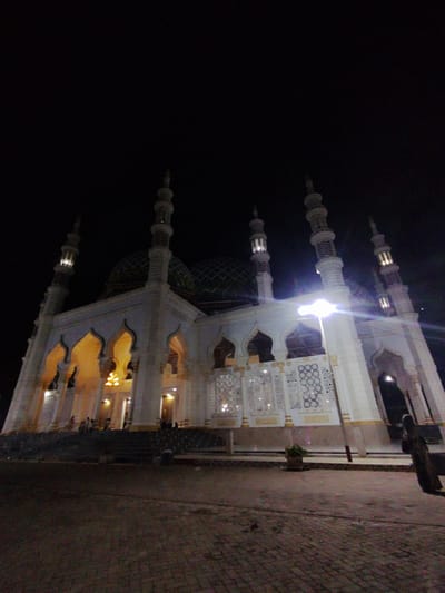 This low-angle nighttime photograph depicts a large, brightly illuminated mosque in Kotapantonlabu, Indonesia. The structure occupies the majority of the frame, contrasting sharply against a uniformly dark, featureless sky. The mosque features a predominantly light-colored, likely white or off-white, facade adorned with multiple tall, slender minarets, each with decorative tiers and internal illumination visible at their pinnacles. At least two large, green, patterned domes with a diamond or scale design are partially visible behind the primary minarets and main building.
The central mosque structure is characterized by multiple grand arched entrances, which emit a warm, yellow light from internal fixtures, including visible chandeliers. Sections of the facade, particularly on the right side, are embellished with white, carved, or perforated screens featuring intricate geometric patterns, such as interlocking circles and stylized star motifs. Robust columns support the main structure.
In the immediate foreground, a dark, paved plaza or courtyard, constructed from brick or paver stones, extends towards a wide, dark staircase that ascends to the mosque's main level. A single, intensely bright street light or floodlight on a pole is positioned in the middle-ground to the right of the mosque, generating a significant lens flare that partially obscures a section of the building's facade. A small potted plant with dark foliage rests on a low ledge near the base of this street light.
On the far right edge of the frame, a lone individual, clad in dark, full-length attire, is observed from behind, standing on the paved ground. Faint, indistinct dark figures are also visible on the mosque's steps and within its illuminated arched doorways, suggesting the presence of other people.