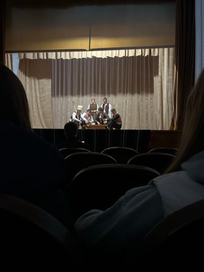 From an audience perspective, the image shows a stage performance in Voiniagovo, Bulgaria. The stage is set with light-colored curtains. Several actors in period costumes are visible on stage. Theater seating is partially visible in the foreground. The image is taken at an angle, and some parts are obscured by the theater seating. The light in the image suggests an indoor location.