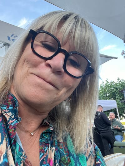 This is a close-up, selfie-style photograph taken outdoors, likely in the city of Saint-Priest-en-Jarez, France, given the visual context. The central subject is a woman with blonde hair and large, dark-rimmed glasses, wearing a colorful, patterned shirt. Her head is tilted, and she is looking downwards, with a slight smile. She is wearing a delicate gold necklace with a single clear pendant. In the background, out of focus, there are other people, white tents or canopies, and green foliage, suggesting an outdoor event or gathering. The sky is visible, with some clouds. One of the background figures appears to be wearing a green jacket and jeans, engaged in conversation. Another figure is wearing a black top and dark pants. The overall environment suggests a daytime setting, possibly in a park or event venue.