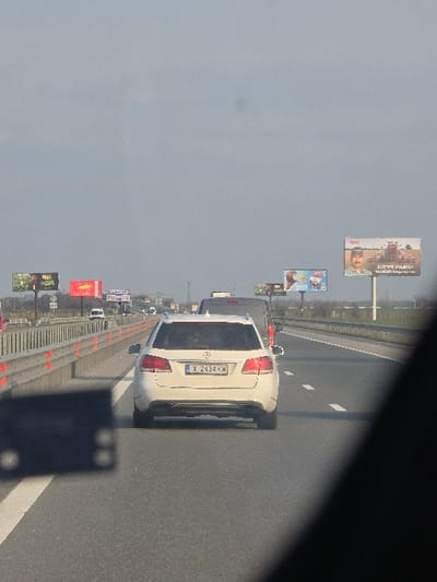 The image displays a daytime highway scene under a predominantly clear, light blue sky, located in the vicinity of Opalchenets, Bulgaria. The perspective is captured from within a vehicle, looking forward into moderate traffic.
In the midground, a white Mercedes-Benz E-Class Estate (wagon) with Bulgarian license plate X 2434 KW occupies the second lane from the right. This vehicle is either moving or stationary within a flow of traffic. Directly behind the Mercedes, a dark-colored van or SUV is partially visible. Additional vehicles are discernible further ahead in all visible lanes.
The highway consists of multiple asphalt lanes marked with white dashed lines. A metal guardrail with orange reflective elements lines the left side of the road. Several large commercial billboards are positioned along the roadside in the distance; one on the right features a man with a mustache, a tractor, and Cyrillic text. An overhead gantry structure is also visible further down the highway. The background horizon shows flat terrain, likely open fields. A dark, out-of-focus structural element of the capturing vehicle partially obstructs the lower-left and a diagonal portion of the lower-right of the frame. No individuals are clearly identifiable.