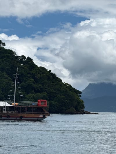 The image captures a coastal scene in Mangaratiba, Brazil. A wooden, multi-deck boat is positioned in the left midground, partially cropped on the left edge. The boat features a brown lower hull, a white upper hull, and a red stripe at the waterline, with black fenders along its side. A tall mast with ladder-like rungs and a decorative white railing shaped like a heart is visible on the upper deck. An orange-red rectangular container, consistent with a life raft or equipment storage, is present on the upper deck. No individuals are clearly discernible on the vessel.
To the right and behind the boat, a densely vegetated, dark green hillside or island shoreline extends into the water. The vegetation consists of numerous tropical trees. The water's edge along the landmass shows exposed rocks. The foreground consists of rippling, grey-blue water.
In the background, distant blue-grey mountains are visible on the right, partially obscured by a layer of white and grey clouds. The sky above displays a mix of clear blue patches and scattered white cumulus clouds, with heavier cloud formations present over the distant mountains. The overall illumination suggests overcast or partly cloudy conditions.