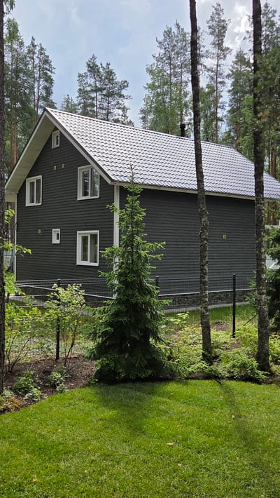 A dark gray, two-story house with a white trim and a gray metal roof sits among tall pine trees in a wooded area. The house has several white-framed windows on both levels. A small, conical evergreen tree stands in the foreground in front of the house, with a neatly mowed green lawn in front of it. A black wire fence with a stone base runs along the property line behind the evergreen tree. The sky is overcast with scattered clouds. The location is identified as the city of Kirillovskoye, Russia. There are no people visible in the image.