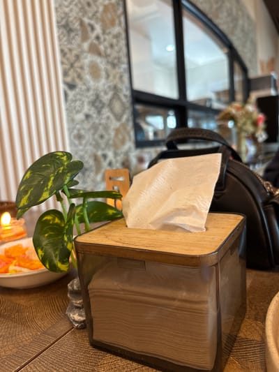 The image captures a close-up of a tissue box with a wooden lid, placed on a wooden table. A portion of a plant with green and yellow leaves is visible to the left of the tissue box. In the background, a black bag and a blurred floral arrangement are discernible. The overall setting appears to be an indoor space, possibly a cafe or restaurant, with patterned wallpaper and a window with black frames. The location is identified as Gajahan, Indonesia. There are no people visible in the image.
