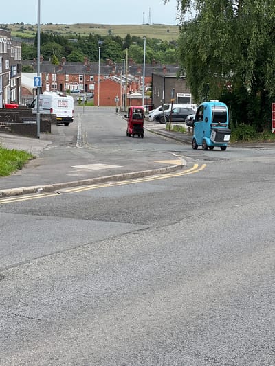 This is a street-level, slightly tilted, eye-level photograph taken in Stoke-on-Trent, United Kingdom. The scene captures a residential street with parked vehicles and houses.
The primary objects in the image are several vehicles. A light blue, three-wheeled vehicle, possibly a small van or autogyro, is parked near the curb. Next to it is a white car, and further down the street, a red van is visible. Two white delivery vans are parked further along the road. One of these vans has text on its side that reads "UK WIDE FOOD SUPPLIES".
The setting appears to be a suburban or semi-rural area, characterized by rows of brick houses with pitched roofs, interspersed with greenery. Trees and bushes line the street and gardens. In the background, a grassy hillside is visible, with power line pylons dotting the landscape. The road itself is asphalt, with yellow double lines indicating parking restrictions.
The time of day appears to be daytime, with overcast or diffused lighting, suggesting a cloudy or possibly early morning/late afternoon setting. No people are clearly visible. The overall impression is of a quiet, everyday scene in a residential neighborhood.