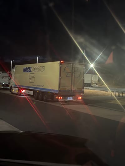 A nighttime outdoor scene captures a yellow semi-trailer truck with "NAGEL-GROUP" written in blue lettering on its side. The truck is parked or moving slowly on an asphalt surface, possibly a highway or rest stop, in Helmstadt, Germany. The background features streetlights casting bright beams and a darker wooded area. The truck has a visible license plate frame, and taillights are illuminated. A smaller white truck is visible in the distance. The image is affected by light flares and reflections.