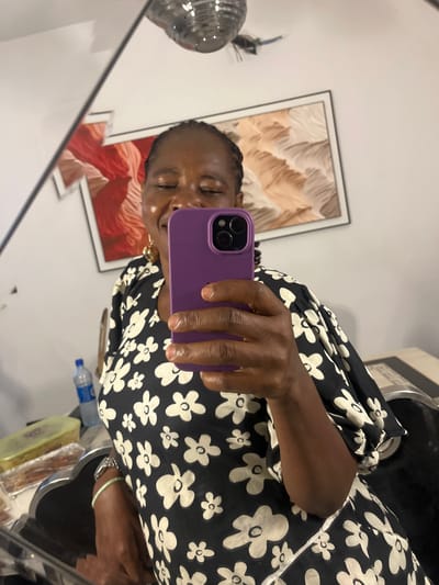 A dark-skinned person with braided hair is taking a selfie in a mirror, holding a purple smartphone. The person is wearing a black and white floral print dress. Behind the person, there is an abstract painting in a frame on the wall. In the foreground, near the person's arm, there is a water bottle, a wrapped food item, and a metal object. The location is inferred to be in Mopo-Akinlade, Nigeria.