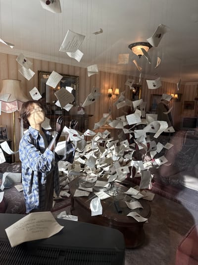 The media file depicts a scene of a room seemingly from the Harry Potter universe, characterized by numerous letters suspended in mid-air. A mannequin dressed in a blue plaid shirt, resembling Harry Potter, is lying on the floor amidst the letters, with a pair of glasses on its face. The room features striped wallpaper, ornate lamps, framed pictures on the walls, and a large wooden table cluttered with more letters and a remote control. The overall impression is of a magical and slightly chaotic event, perhaps the famous "Hogwarts letters" scene from the books. The lighting suggests daytime, with sunlight casting shadows on the floor and furniture. Several letters prominently display the address "MR H POTTER," "The Cupboard under the Stairs," "1 Privet Drive," "Little Whinging," and "SURREY," confirming the iconic Harry Potter theme. The scene appears to be a display or exhibit, possibly in a museum or themed attraction, given the mannequin and the meticulous arrangement of the letters.