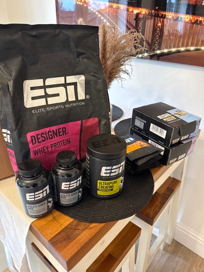 The scene captures a static arrangement of Elite Sports Nutrition (ESI) products on a wooden table, indicative of an interior setting in Mid Suffolk, United Kingdom. Prominently displayed is a large black pouch of "DESIGNER. WHEY PROTEIN" featuring a pink graphic. Adjacent are two black bottles labeled "ATHLETE STACK: MEN" and a black tub of "ULTRAPURE CREATINE PREMIUM GRADE" with a yellow label. To the right, three rectangular black boxes, one visibly marked "PROTEIN BAR," are stacked alongside several smaller black sachets, some with orange-toned details. The products are positioned on a light brown wooden table, partially covered by a white fabric on the left and a circular black woven placemat. In the background, a white wall displays a large framed print depicting a brightly lit bridge at night, characterized by numerous warm-toned lights. A decorative element of dried, feathery brown plant material is also visible. No individuals are present.
