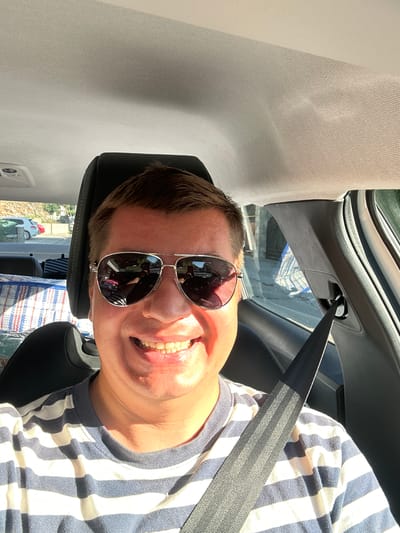 A man wearing sunglasses and a striped t-shirt is sitting in the driver's seat of a car, taking a selfie. The car interior is visible, including the dashboard, car seat, and seatbelt. Outside the car, through the windows, a street scene is visible with buildings and parked cars. The sunlight suggests it is daytime. The man is smiling broadly. The location is identified as Provodina, Montenegro.