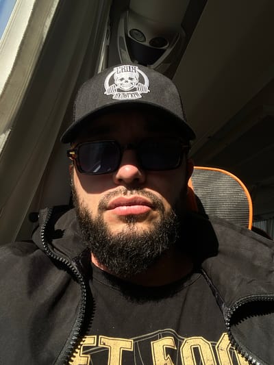 This is a close-up, vertically oriented selfie of a man wearing a black baseball cap with a skull logo and sunglasses. He has a beard and is wearing a black jacket over a black t-shirt with gold lettering. The lighting suggests it is daytime, with strong sunlight casting shadows. The background is blurred, but hints of an interior space are visible, possibly a vehicle or room with vents and a window. The man appears to be looking slightly upwards and to the side. The overall mood is casual and perhaps thoughtful. Visible text on the cap reads "GRIND" and on the t-shirt appears to be part of a logo or phrase in gold lettering. The location is identified as Vitino, Russia.