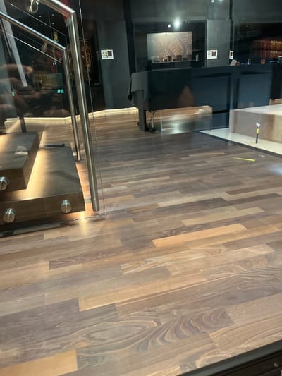 This is a tilted, low-angle, eye-level, full shot of a staircase and a wooden floor in what appears to be a modern building. The staircase has glass sides and a metallic handrail. The steps are made of dark wood and are illuminated from underneath, casting a warm glow. The floor is made of long, narrow wooden planks with a natural grain pattern, varying in shades of brown and grey.
In the background, there are reflections in the glass. One reflection shows what looks like a wall with some framed items and a display cabinet. Further in the background, books are visible on shelves. Another reflection shows a person taking the photo, with dark hair and wearing dark clothing, holding a phone.
There is a yellow object on the floor near the top edge of the image, and a small white electronic device with a few buttons on a dark surface in the background, possibly a control panel.
The lighting is artificial, with the illuminated staircase and the overall tone suggesting it could be evening or indoors. The location cues are minimal, but the scene appears to be an interior space, possibly a museum, library, or upscale commercial building. No specific activity or event is clearly depicted beyond the implied presence of people navigating the space. The mood is neutral and clean, with an emphasis on the modern design and materials.