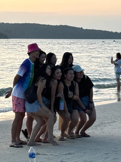Eight individuals, comprising one male and seven females, are posing for a photograph on the sandy shore of Cenang Beach, Malaysia, during what appears to be late afternoon or early evening. The male, positioned on the far left, wears a pink bucket hat, a multi-colored (blue, purple, and white) short-sleeved shirt, and pink shorts with a shark pattern, along with black flip-flops. He is smiling and has his left arm around a female next to him. The seven females, predominantly of Asian ethnicity, are mostly dressed in dark tops and denim shorts. One female wears a light blue baseball cap, and another holds a clear plastic cup with a straw. They are arranged in a compact group, smiling and looking towards an unseen camera. Several individuals in this group are barefoot, while others wear flip-flops. In the right background, a separate female individual stands knee-deep in the ocean, facing the posing group and holding a smartphone with both hands as if taking a photograph. The environment includes calm ocean water with minimal waves, a distant forested landmass on the horizon, and a sky exhibiting soft orange, pink, and grey hues. A clear plastic water bottle is visible on the sand in the lower foreground.