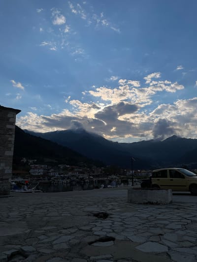 A westward-facing, vertically-oriented photograph captures a scene from Skala Potamias, Greece. The composition is divided into three sections. The left portion showcases a blue sky with scattered white clouds, some illuminated by sunlight. The central area features a dark, silhouetted mountain range partially obscured by clouds. To the right, a portion of the city is visible, including buildings and a parked yellow car. The foreground includes a stone structure and paved walkway, suggesting an elevated viewing point.