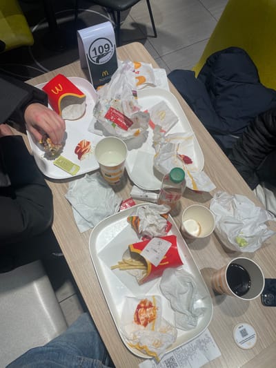 A cluttered table at McDonald's holds used trays filled with remnants of a meal: crumpled burger wrappers, empty fry containers with some fries still inside, ketchup packets, used cups, and a half-eaten burger in someone's hands. A drink, a bottle of water, receipts, a phone, and a table number are also visible, suggesting a group or individual has recently finished eating. A jacket sits on a chair beside the table, indicating someone is still present.